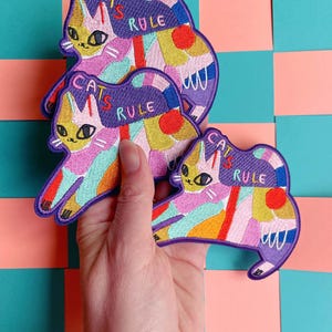 Può includere: Tre toppe ricamate colorate a forma di gatto. Ogni toppa presenta un disegno di gatto con le parole "CATS RULE" in uno stendardo viola. Il disegno del gatto include colori giallo, rosa, blu e arancione.