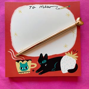 May include: A red notepad with the text "To new" and a gold pen with a heart-shaped top. The notepad features illustrations of a black cat, a cup of coffee, and a white strawberry.