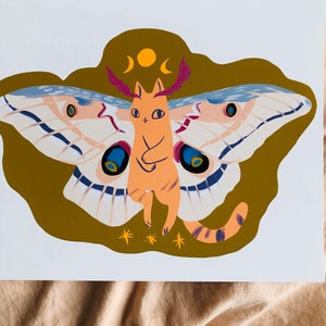May include: A whimsical illustration of a cat with moth wings. The cat is orange with a pink nose and is standing on a gold background with a crescent moon and a sun above it. The moth wings are blue, white, and pink with black spots.