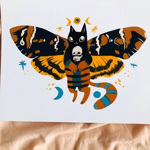 May include: A black cat with a skull in its paws, wearing a moth costume with yellow, orange, and black wings. The cat is surrounded by stars and crescent moons.