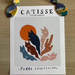 May include: A white print with the text "CATISSE THE CAT GHOSTS" and ".PURRS COLLECTION."  The print features a colorful illustration of five cats in a ghost-like form with a large orange sun in the background.