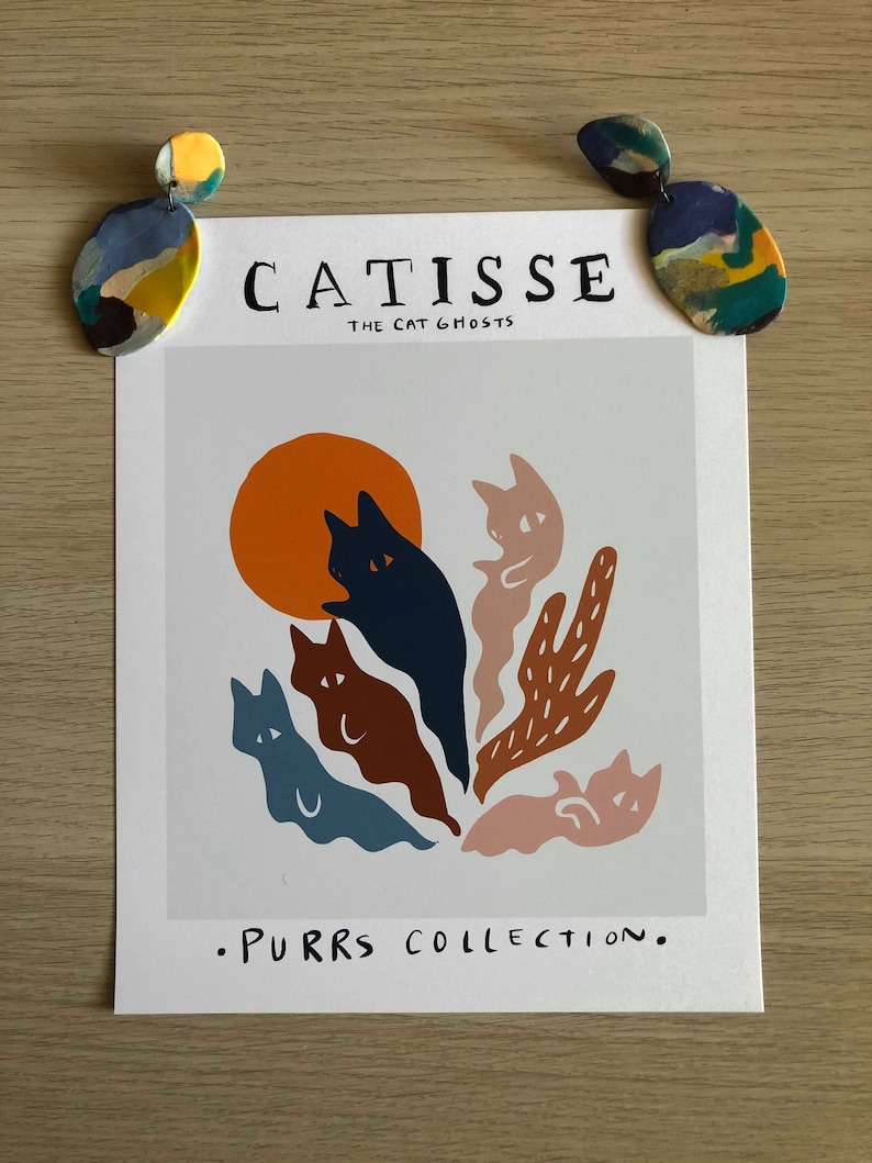 May include: A white print with the text "CATISSE THE CAT GHOSTS" and ".PURRS COLLECTION."  The print features a colorful illustration of five cats in a ghost-like form with a large orange sun in the background.