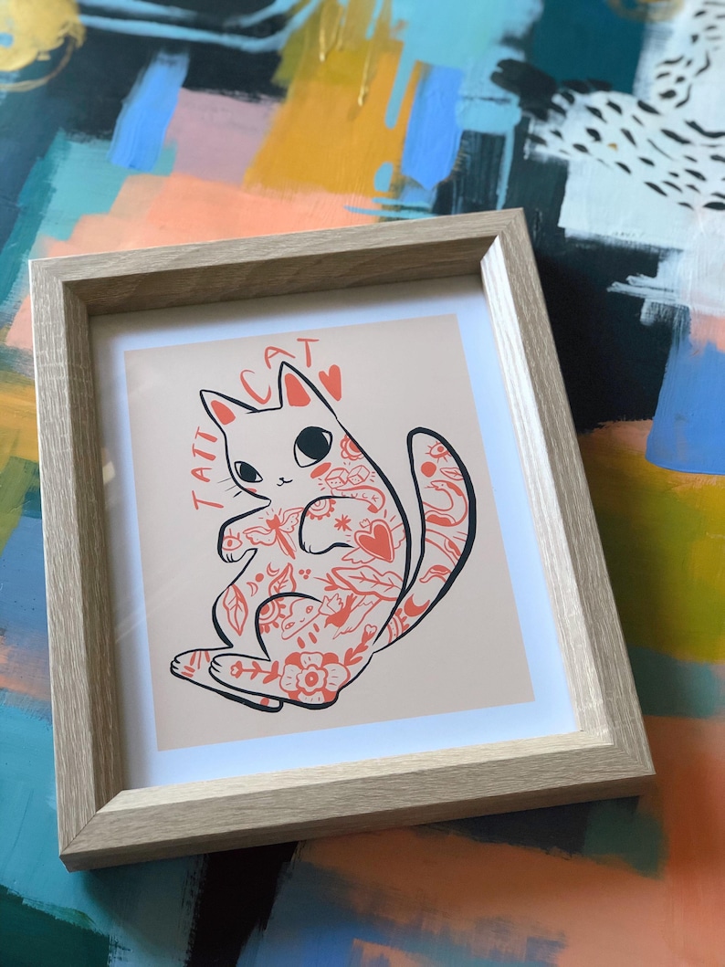 Puede incluir: Una impresi&oacute;n enmarcada de un gato blanco con tatuajes rojos. El gato est&aacute; enroscado con la cola envuelta alrededor de su cuerpo. La impresi&oacute;n tiene el texto "TATT CAT" en letras negras.