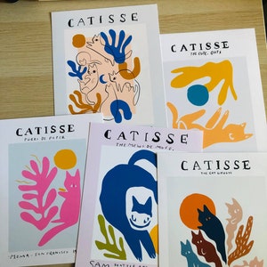 May include: Five colorful prints with the title "CATISSE" and different subtitles. The prints feature abstract cat illustrations with bold colors and simple lines. The prints are displayed on a wooden surface.