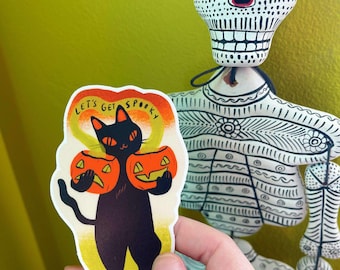 May include: A black cat holding two orange jack-o'-lantern pumpkins with the text "Let's get spooky" in a yellow, orange, and red circle.