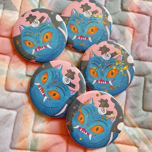 May include: Five round buttons with a whimsical cat design. The cat has blue fur, orange eyes, and white fangs. The background is pink and gray with a black hat and gold glitter accents. The buttons are arranged on a quilted fabric.