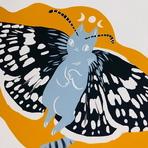 May include: A blue cat with black and white butterfly wings against a yellow background. The cat has a crescent moon on its head.
