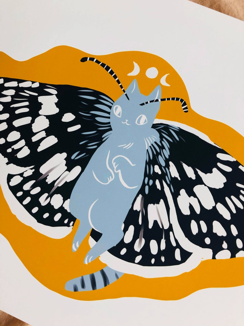 May include: A blue cat with black and white butterfly wings against a yellow background. The cat has a crescent moon on its head.