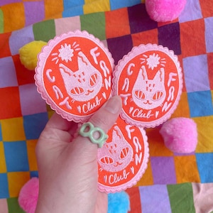 May include: Three orange and pink embroidered patches with a cat design and the text "Cat Fan Club". A green ring is worn on a finger. The patches are on a colorful, patterned background with pink, yellow, and blue pom-poms.