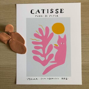 May include: A pink cat with a yellow sun and a pink leaf design on a light blue background. The text "CATISSE PURRS DE PAPER" is at the top of the print. The text ".MOMA - SAN FRANCISCO 1982." is at the bottom of the print.