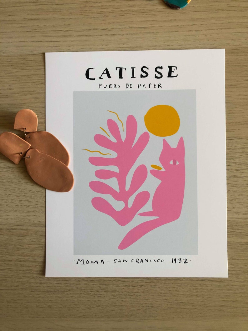 May include: A pink cat with a yellow sun and a pink leaf design on a light blue background. The text "CATISSE PURRS DE PAPER" is at the top of the print. The text ".MOMA - SAN FRANCISCO 1982." is at the bottom of the print.