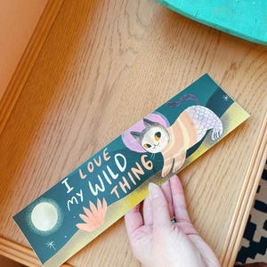 May include: A bookmark with the text "I LOVE MY WILD THING" and an illustration of a cat. The cat is orange and white with a pink head covering. The background is dark teal with a moon and a flower.