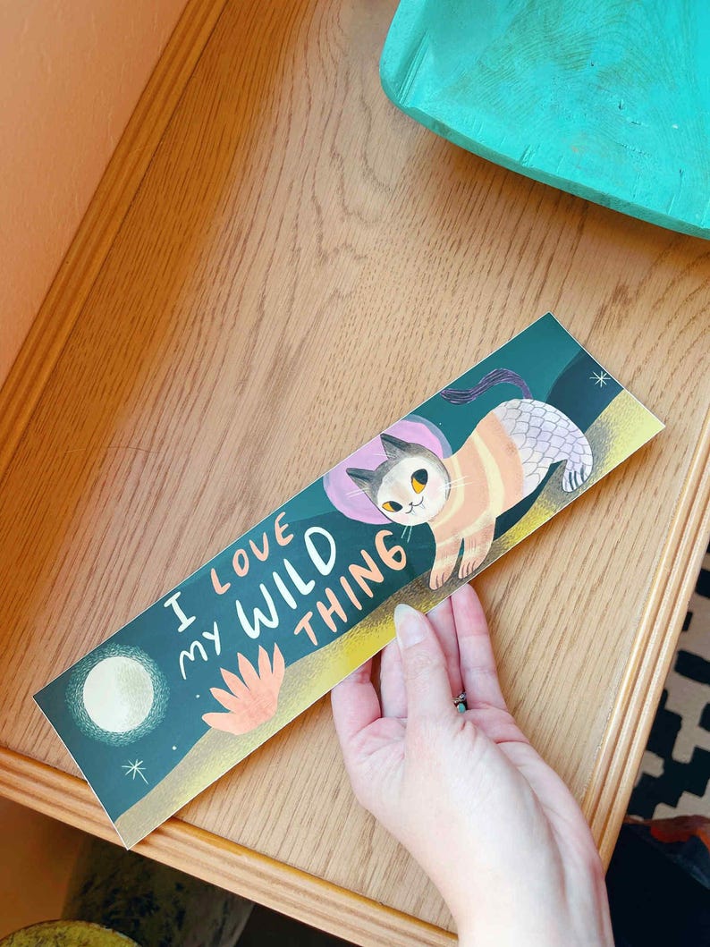 May include: A bookmark with the text "I LOVE MY WILD THING" and an illustration of a cat. The cat is orange and white with a pink head covering. The background is dark teal with a moon and a flower.