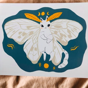 May include: A white cat with butterfly wings and a moon and stars design. The cat has a yellow crescent moon on its head and is surrounded by a teal blue background.