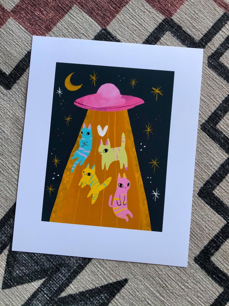 May include: A print of a cartoon illustration of four cats being abducted by a flying saucer. The cats are pink, yellow, blue, and green. The saucer is pink and has a beam of light shining down on the cats. The background is a dark blue with white stars.