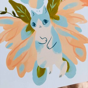 May include: A white cat with blue and orange wings. The cat has a whimsical expression and is surrounded by green leaves. The wings are painted in a watercolor style with soft, blended colors.