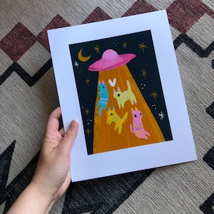 May include: A pink flying saucer with a yellow beam of light shining down on four cats. The cats are different colors: blue, yellow, orange, and pink. The background is a dark blue with white stars.