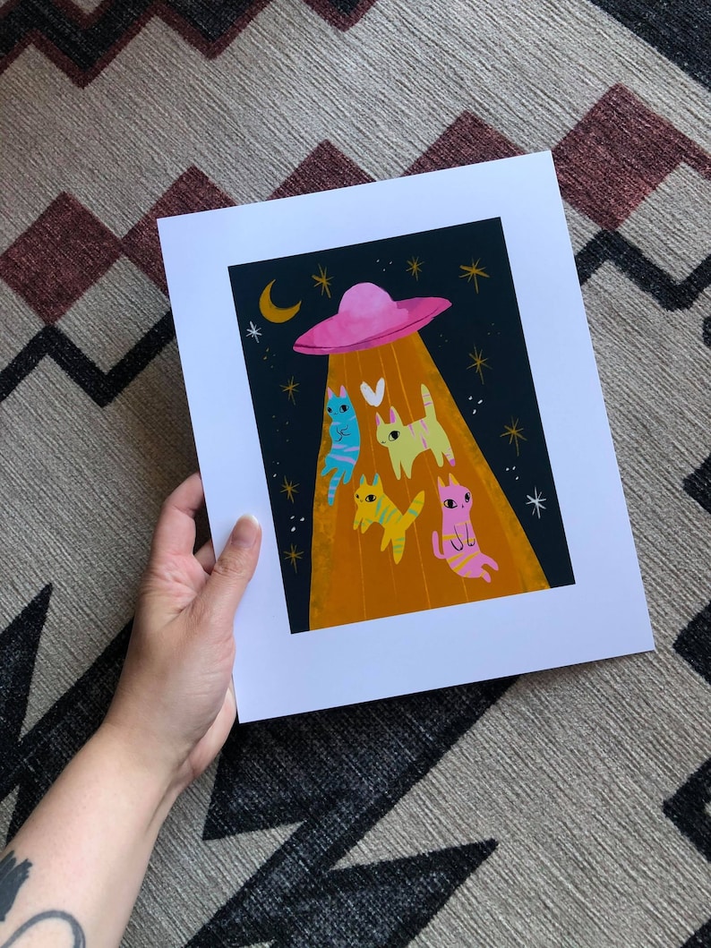 May include: A pink flying saucer with a yellow beam of light shining down on four cats. The cats are different colors: blue, yellow, orange, and pink. The background is a dark blue with white stars.