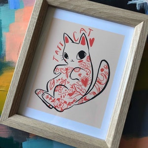 Puede incluir: Una impresi&oacute;n enmarcada de un gato blanco con tatuajes rojos. El gato est&aacute; sentado sobre un fondo rosa claro. El texto "TATT CAT" est&aacute; escrito en rojo sobre el gato.