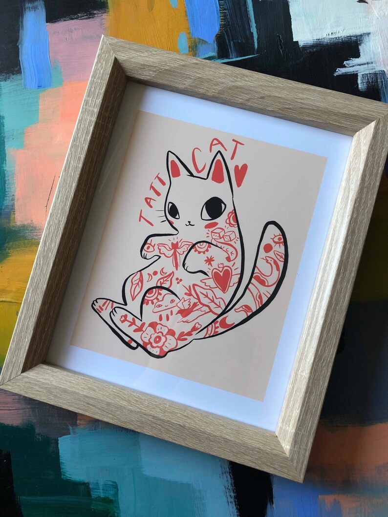 Puede incluir: Una impresi&oacute;n enmarcada de un gato blanco con tatuajes rojos. El gato est&aacute; sentado sobre un fondo rosa claro. El texto "TATT CAT" est&aacute; escrito en rojo sobre el gato.