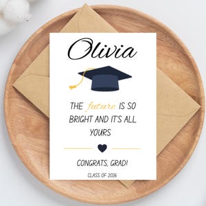 May include: A graduation card with the name "Olivia" in elegant script. The card features a graduation cap illustration and the text "THE future IS SO BRIGHT AND IT'S ALL YOURS CONGRATS, GRAD! CLASS OF 2026". The card is on a wooden tray.