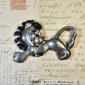 May include: A silver-toned metal brooch shaped like a stylized lion in a leaping pose. The lion features black enamel detailing on the mane and tail, with small red enamel accents on the mouth and paws, and a single clear rhinestone for the eye.