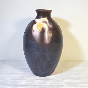 May include: A dark purple ceramic vase with a smooth, rounded body and a narrow neck. The upper portion features a raised, three-dimensional white and yellow flower relief that blends into the dark purple glaze of the vessel's exterior surface.