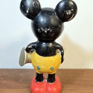 May include: Vintage Mickey Mouse figurine, featuring a black head and ears, yellow shorts, and red shoes. The figurine has the text "Mickey Mouse Walt Disney" on the back. The figure is standing on a wooden surface.