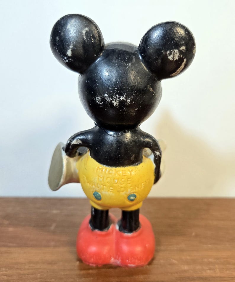 May include: Vintage Mickey Mouse figurine, featuring a black head and ears, yellow shorts, and red shoes. The figurine has the text "Mickey Mouse Walt Disney" on the back. The figure is standing on a wooden surface.