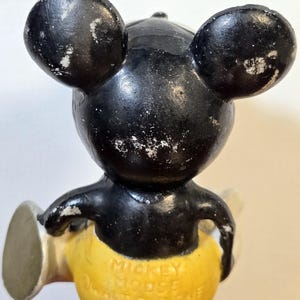 May include: Vintage Mickey Mouse figurine with black head and ears, yellow shorts, and white gloves. The figurine has visible wear and tear, with the text "Mickey Mouse" and "Walt Disney" on the shorts. A classic collectible.