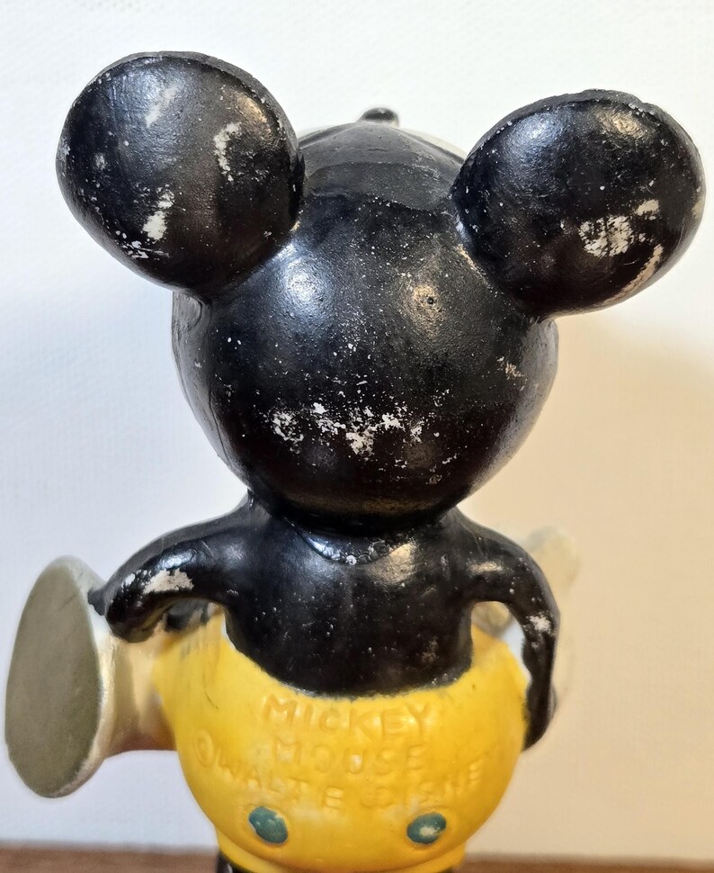 May include: Vintage Mickey Mouse figurine with black head and ears, yellow shorts, and white gloves. The figurine has visible wear and tear, with the text "Mickey Mouse" and "Walt Disney" on the shorts. A classic collectible.