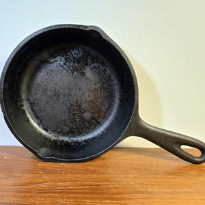 May include: A black cast iron frying pan with a long handle and a hanging hole. The pan sits on a wooden surface. The interior of the pan has a textured surface.
