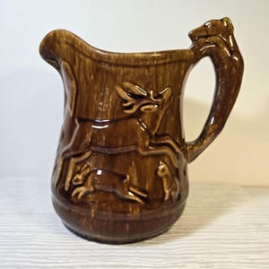 Puede incluir: Una jarra de cerámica marrón con asa y boquilla. La jarra presenta imágenes en relieve de un ciervo, conejos y un animal pequeño. El asa está adornada con un oso. La jarra tiene un acabado brillante.