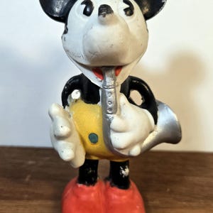 May include: A vintage Mickey Mouse figurine playing a silver saxophone. The figure has a white face, black ears, and is wearing a yellow shirt with a blue button, black shorts, and red shoes. The figurine is approximately 4 inches tall.