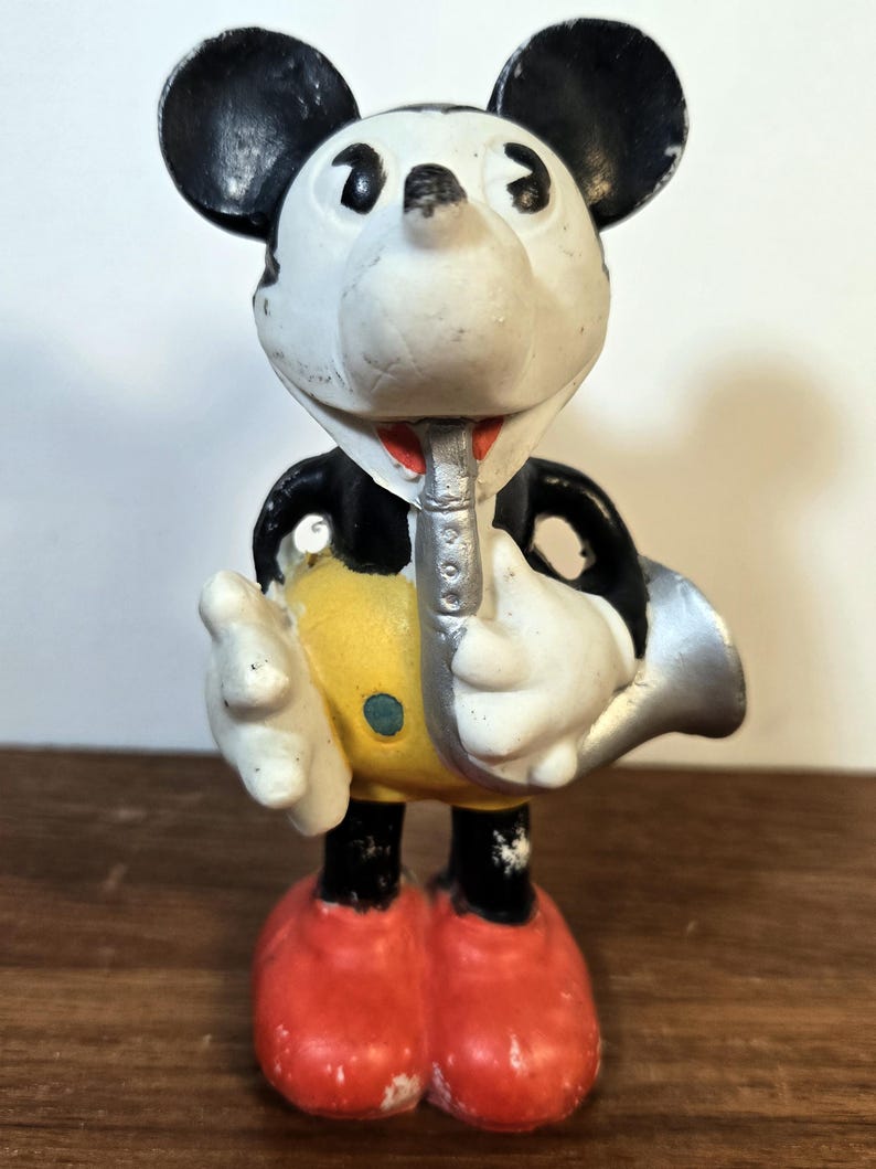 May include: A vintage Mickey Mouse figurine playing a silver saxophone. The figure has a white face, black ears, and is wearing a yellow shirt with a blue button, black shorts, and red shoes. The figurine is approximately 4 inches tall.