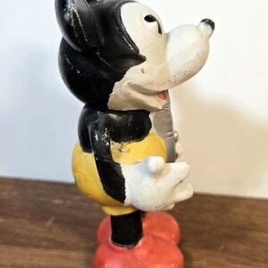 May include: Vintage Mickey Mouse figurine in a classic pose. The figure features a black head and ears, a white face and hands, a yellow torso, black shorts, and red shoes. The figurine is standing on a wooden surface.
