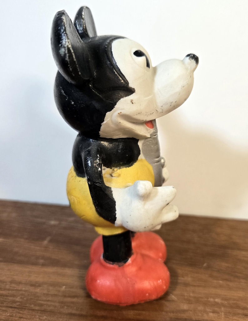 May include: Vintage Mickey Mouse figurine in a classic pose. The figure features a black head and ears, a white face and hands, a yellow torso, black shorts, and red shoes. The figurine is standing on a wooden surface.