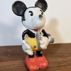May include: Vintage Mickey Mouse figurine in a tuxedo, playing a silver saxophone. The figure has black ears, a white face, and red shoes. The yellow vest and black tuxedo are visible.
