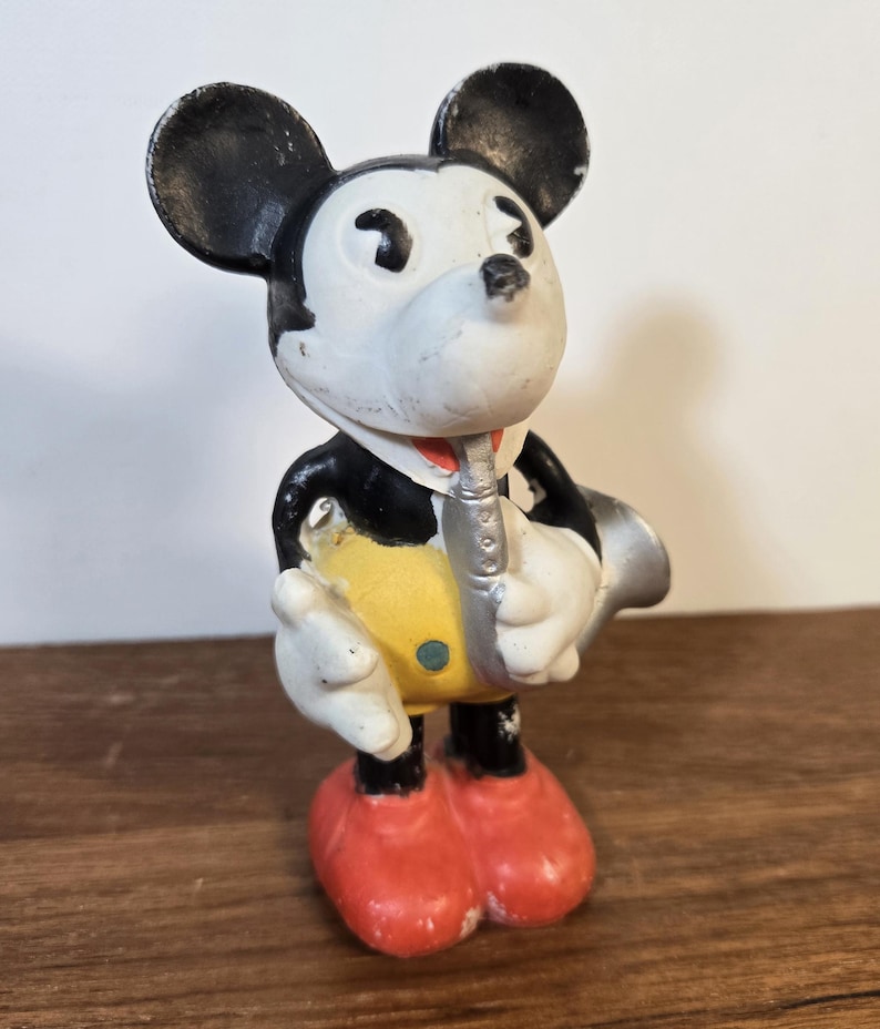 May include: Vintage Mickey Mouse figurine in a tuxedo, playing a silver saxophone. The figure has black ears, a white face, and red shoes. The yellow vest and black tuxedo are visible.