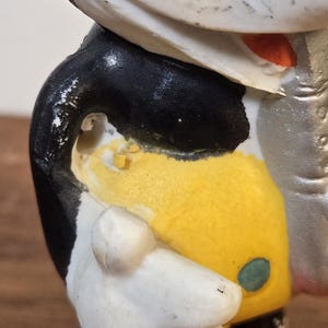 May include: A vintage figurine of a cartoon character, featuring a white face, black jacket, yellow torso, and white gloves. The figure has a red nose and a green button detail. The paint shows signs of wear, indicating its age.