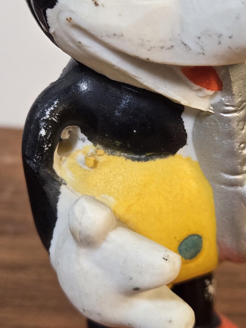 May include: A vintage figurine of a cartoon character, featuring a white face, black jacket, yellow torso, and white gloves. The figure has a red nose and a green button detail. The paint shows signs of wear, indicating its age.