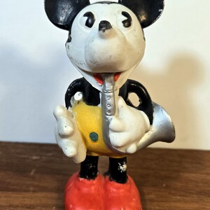 May include: Vintage Mickey Mouse figurine playing a silver saxophone. The figure has black ears, a white face, and a red mouth. Mickey wears a black jacket, yellow shorts, red shoes, and white gloves. The figurine is standing on a wooden surface.