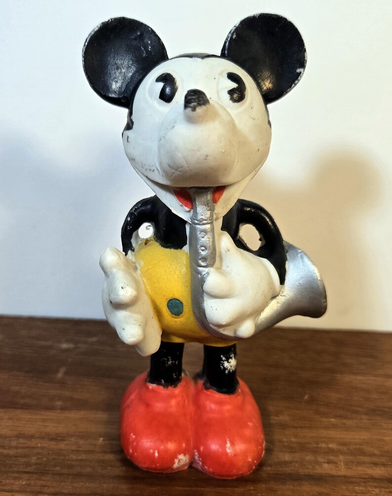 May include: Vintage Mickey Mouse figurine playing a silver saxophone. The figure has black ears, a white face, and a red mouth. Mickey wears a black jacket, yellow shorts, red shoes, and white gloves. The figurine is standing on a wooden surface.