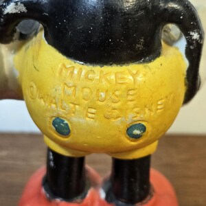 May include: Vintage Mickey Mouse figurine with black head and arms, yellow shorts, and red shoes. The text "MICKEY MOUSE" and "WALT E. DISNEY" are embossed on the shorts. The figurine has a distressed, aged appearance.