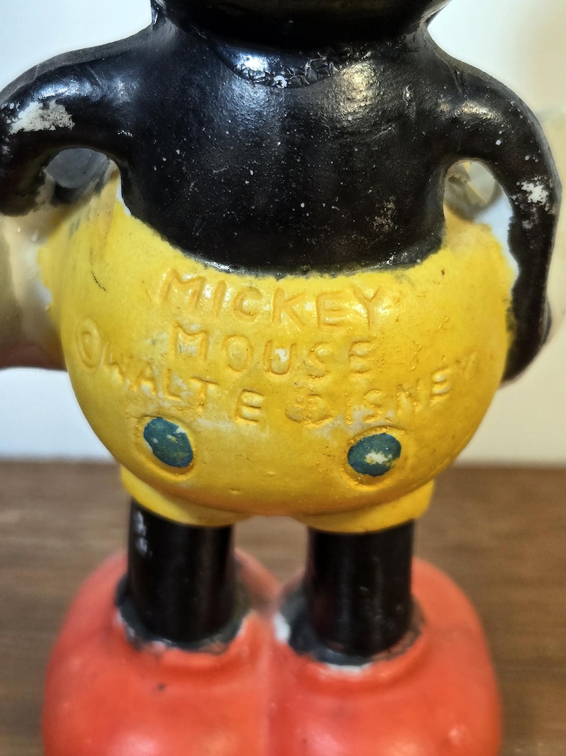 May include: Vintage Mickey Mouse figurine with black head and arms, yellow shorts, and red shoes. The text "MICKEY MOUSE" and "WALT E. DISNEY" are embossed on the shorts. The figurine has a distressed, aged appearance.