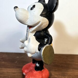 May include: Vintage Mickey Mouse figurine in a standing pose. The figure has a white face, black ears and body, red shorts, and yellow shoes. The figurine is holding a silver object and has a gold-colored disc on its back.