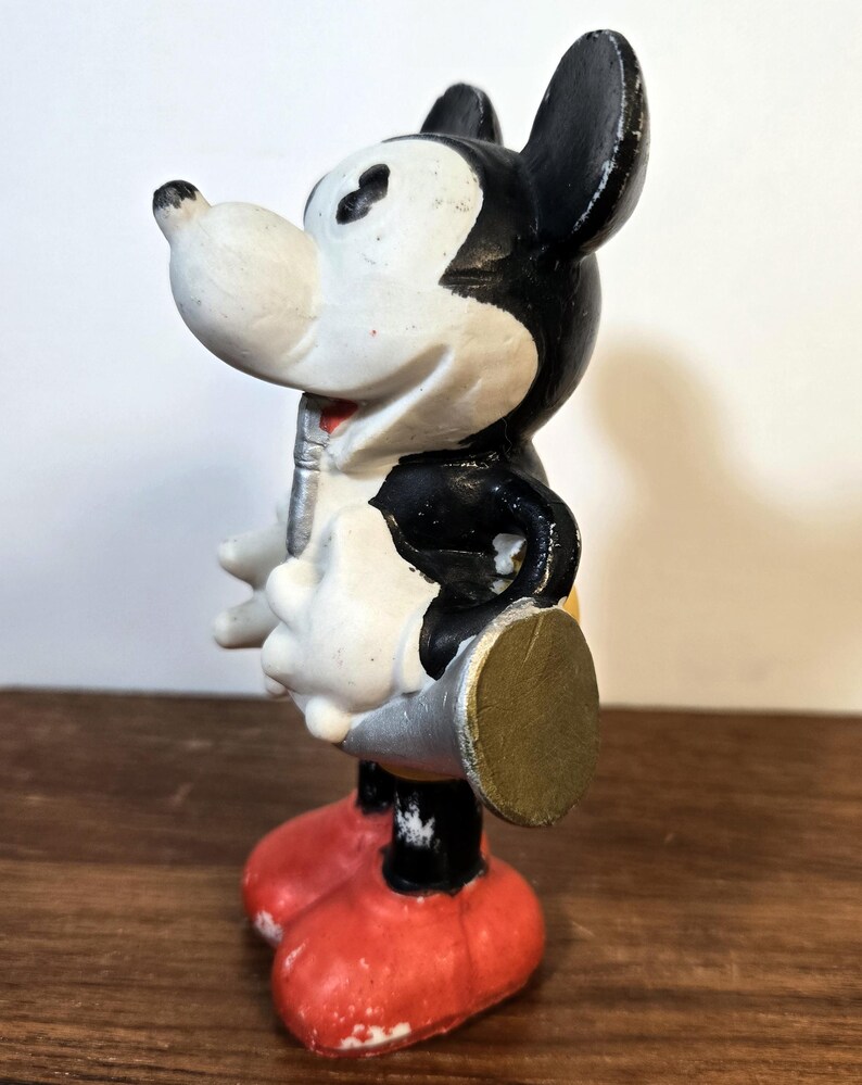 May include: Vintage Mickey Mouse figurine in a standing pose. The figure has a white face, black ears and body, red shorts, and yellow shoes. The figurine is holding a silver object and has a gold-colored disc on its back.