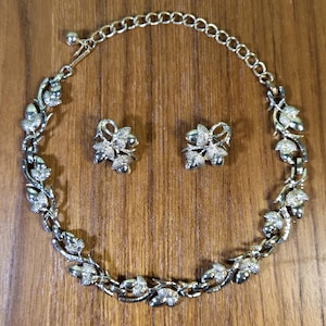 May include: A silver-toned jewelry set featuring a necklace and matching earrings. The necklace and earrings are adorned with a leaf and berry design. The necklace has a chain closure. The set is displayed on a wooden surface.