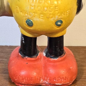 May include: A vintage Mickey Mouse figurine, featuring a yellow torso, black legs, and red shoes. The text "MICKEY MOUSE &copy; WALT E. DISNEY" is visible on the torso. The figurine is marked "MADE IN JAPAN" on the bottom.
