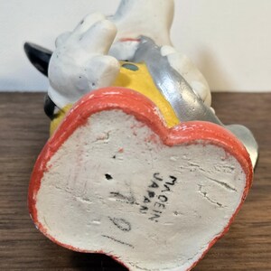 May include: A vintage figurine of a cartoon character, featuring a white body, black ears, and a yellow and gray outfit. The base is red and the bottom reads "MADE IN JAPAN".